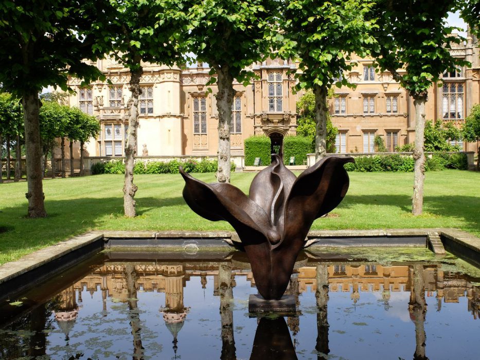 A large sculpture by Anne Curry shown in the middle of a pond in front of a stately home
