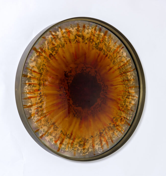 The circular Baltic Iris Mirror in metal frame with brass finish by Tom Palmer studio, the amber and orange colour of the cast lens inspired by the rich tones of amber found around the Baltic Sea.